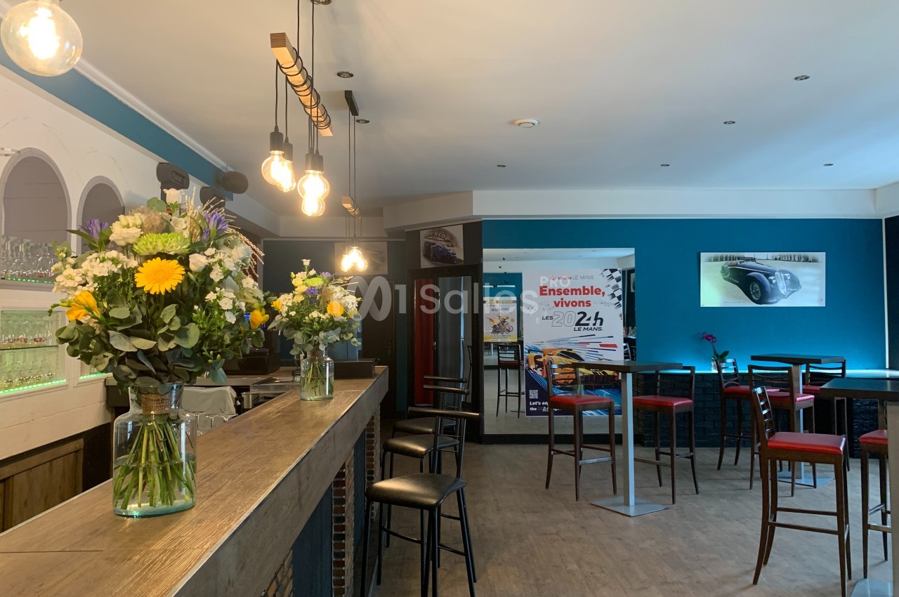 Intérieur d'un café moderne avec bar en bois, tabourets, tables hautes et bouquets de fleurs colorées.