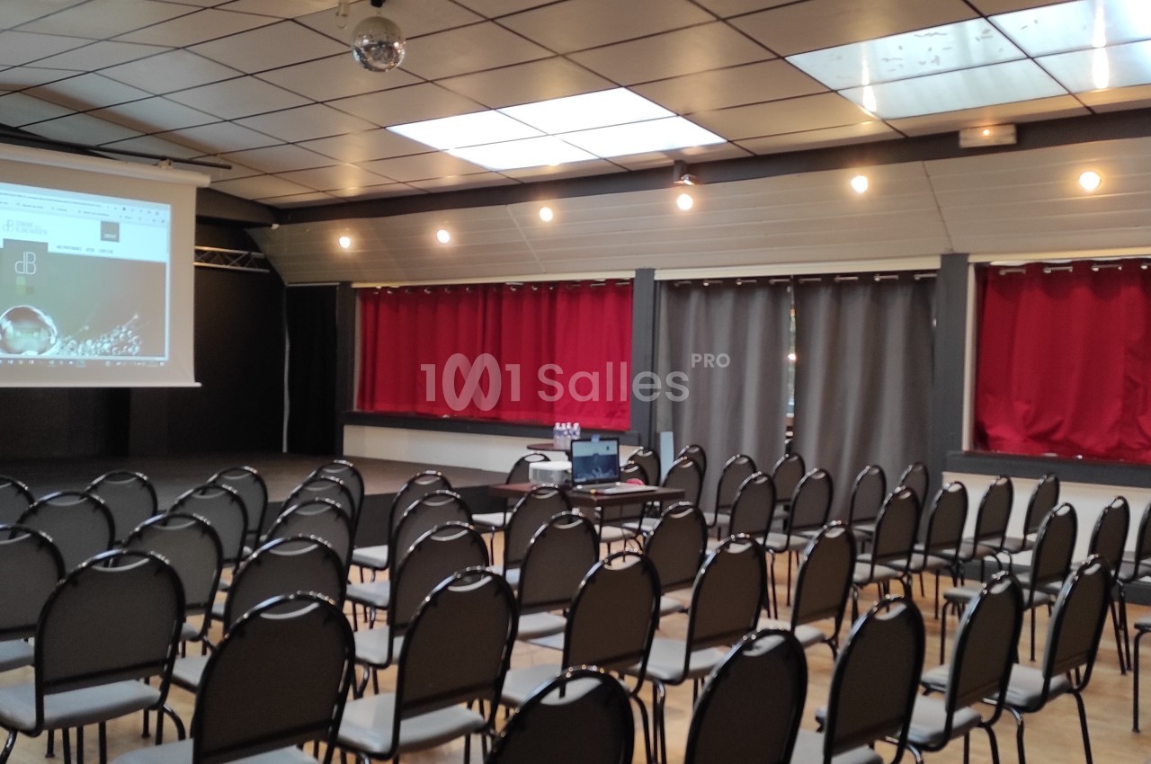 Salle de conférence avec des rangées de chaises vides, un écran de projection allumé et des rideaux rouges en arrière-plan.