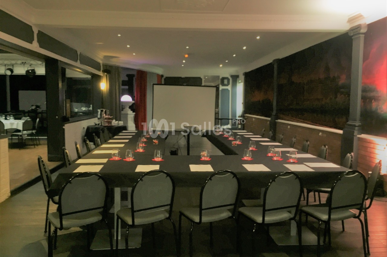 Salle de réunion avec tables disposées en U, chaises alignées, tableau blanc et éclairage tamisé.