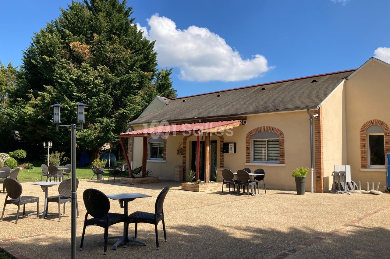 Terrasse avec tables et chaises devant un bâtiment beige à toit en ardoise, entouré de végétation.