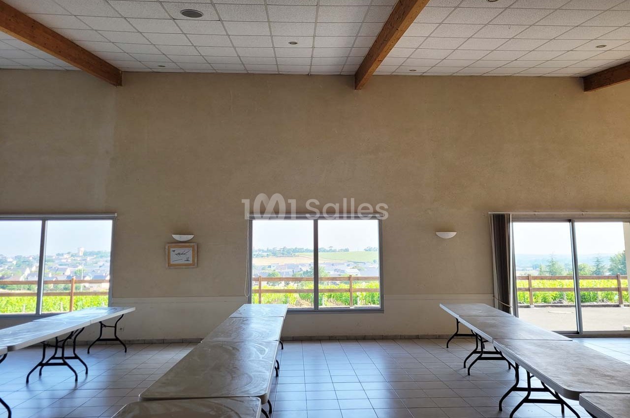 Salle lumineuse avec grandes fenêtres, tables pliantes alignées et vue dégagée sur un paysage extérieur.