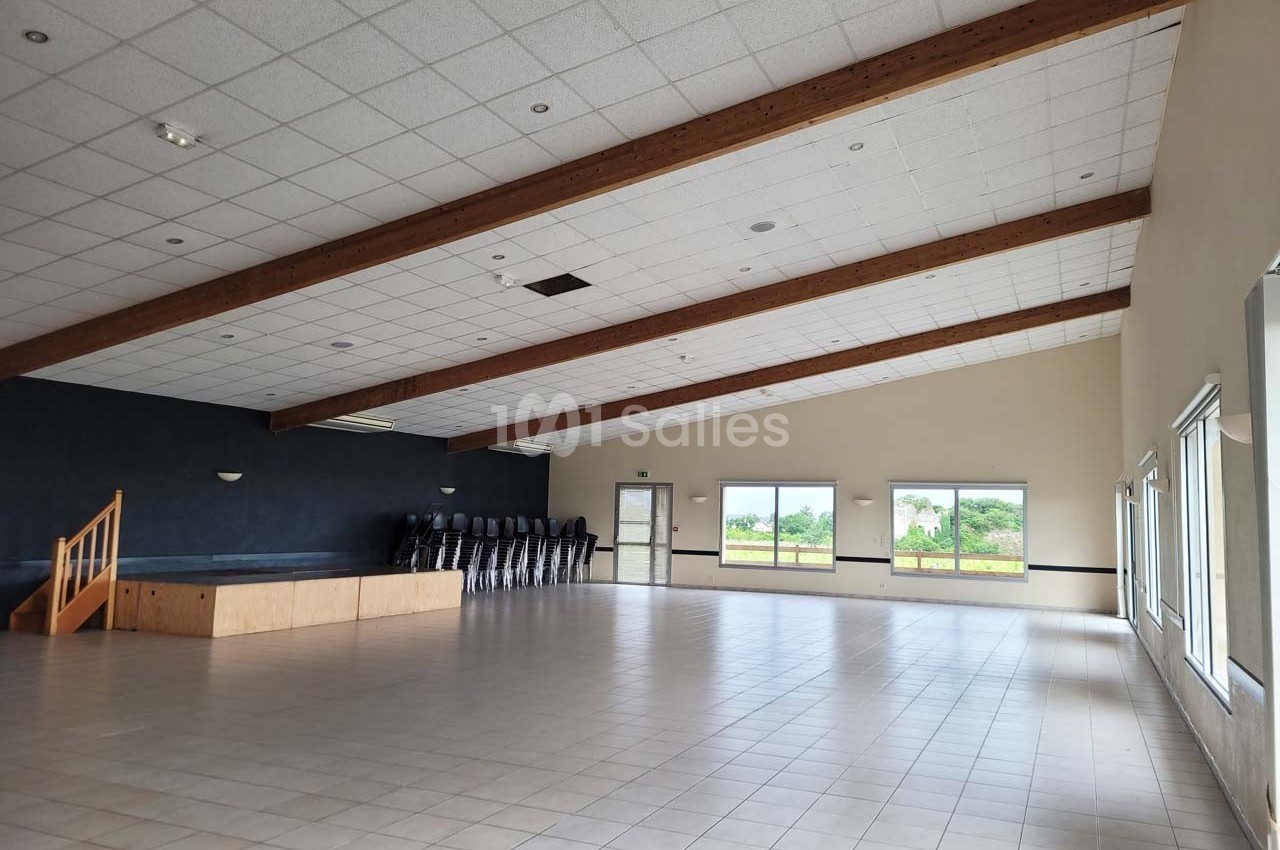 Salle spacieuse avec sol carrelé, scène en bois, chaises empilées et grandes fenêtres donnant sur un paysage verdoyant.