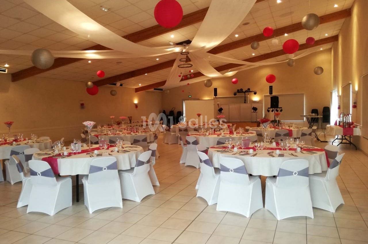 Salle de réception décorée avec des tables rondes dressées, des nappes blanches et des suspensions rouges et grises.