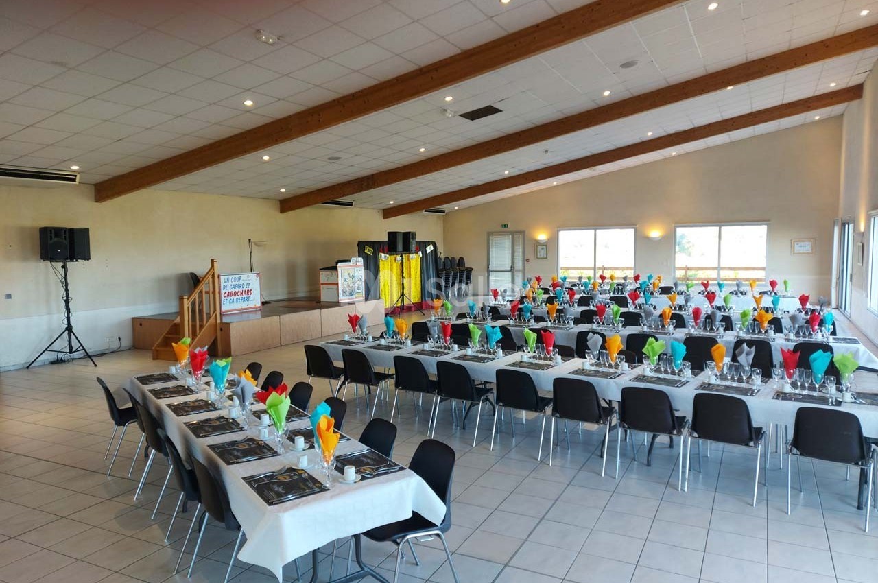 Salle de réception décorée avec des tables alignées, nappes blanches et serviettes colorées, prête pour un événement.
