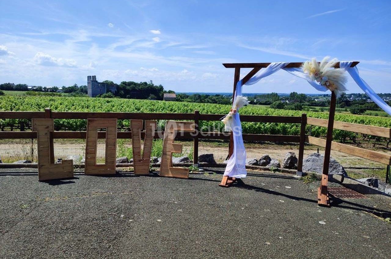 Structure en bois formant le mot ’LOVE’ et arche décorée de tissu blanc, devant un vignoble sous un ciel dégagé.