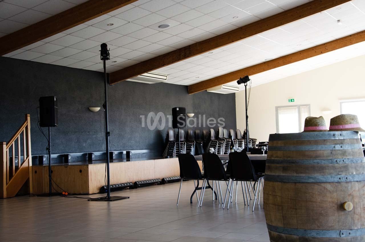 Salle vide avec scène, chaises empilées, tables noires, tonneau en bois au premier plan et éclairage naturel.