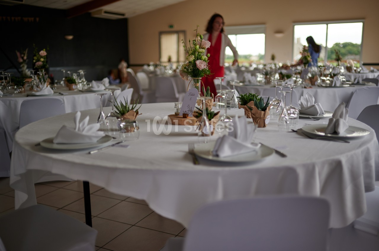 Salle de réception avec tables rondes dressées, nappes blanches, décorations florales et lumière naturelle.