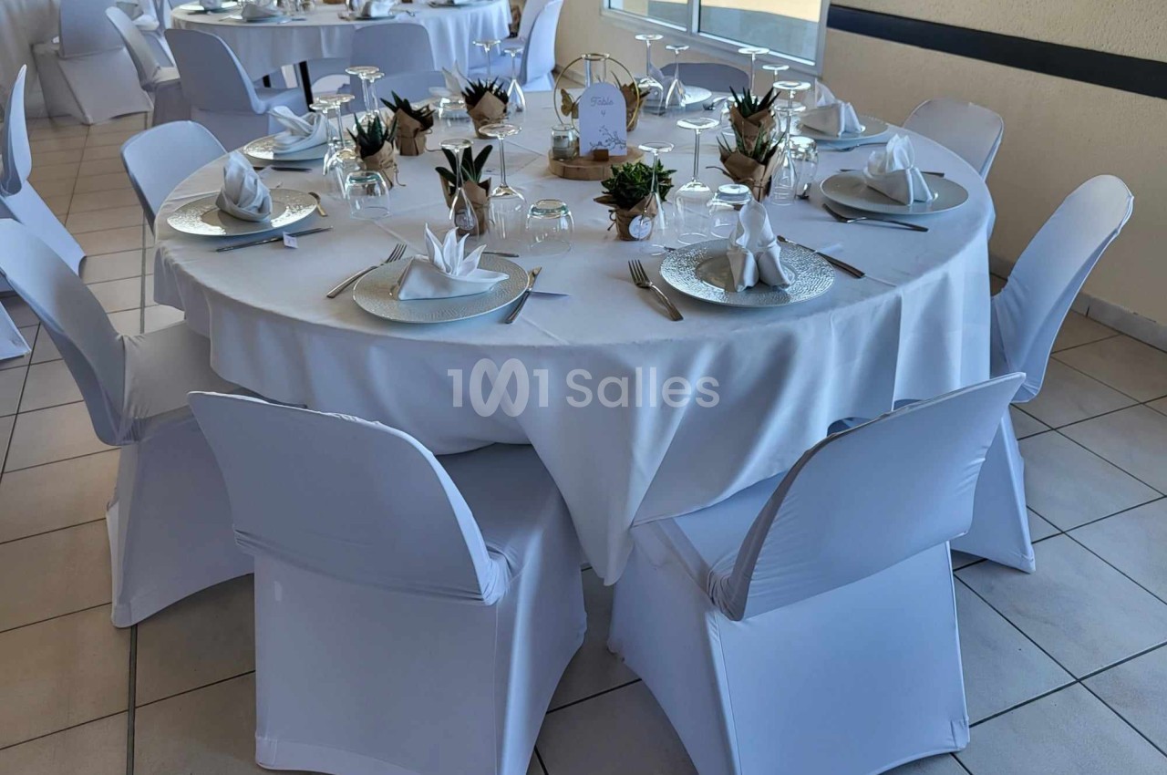 Table ronde décorée pour un événement, avec nappes blanches, serviettes pliées, vaisselle élégante et petites plantes.