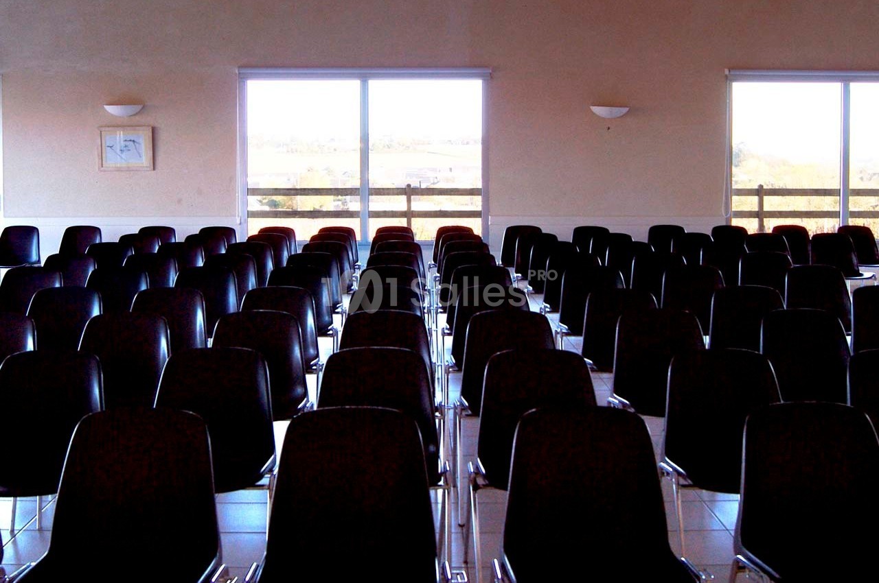 Salle avec des rangées de chaises noires alignées, éclairée par la lumière naturelle provenant de grandes fenêtres.