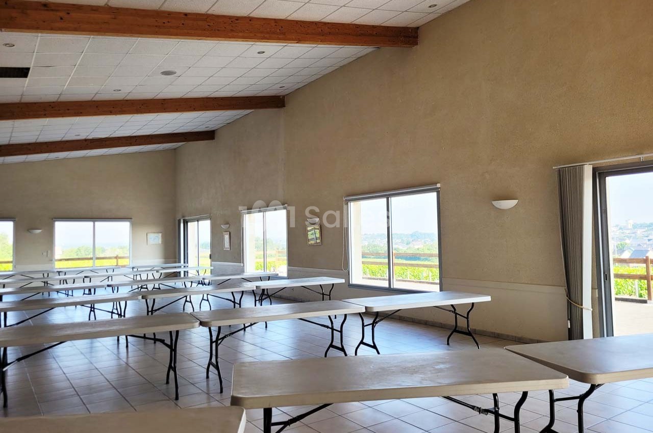 Salle lumineuse avec de grandes fenêtres, tables pliantes alignées et plafond avec poutres apparentes.