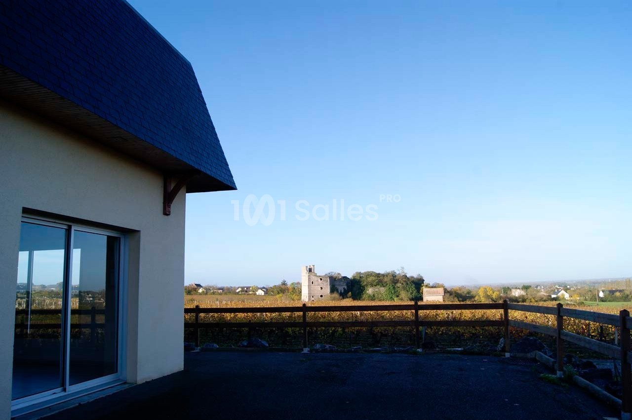 Vue d'un paysage rural avec un bâtiment moderne à gauche, des vignes et un château au loin sous un ciel dégagé.