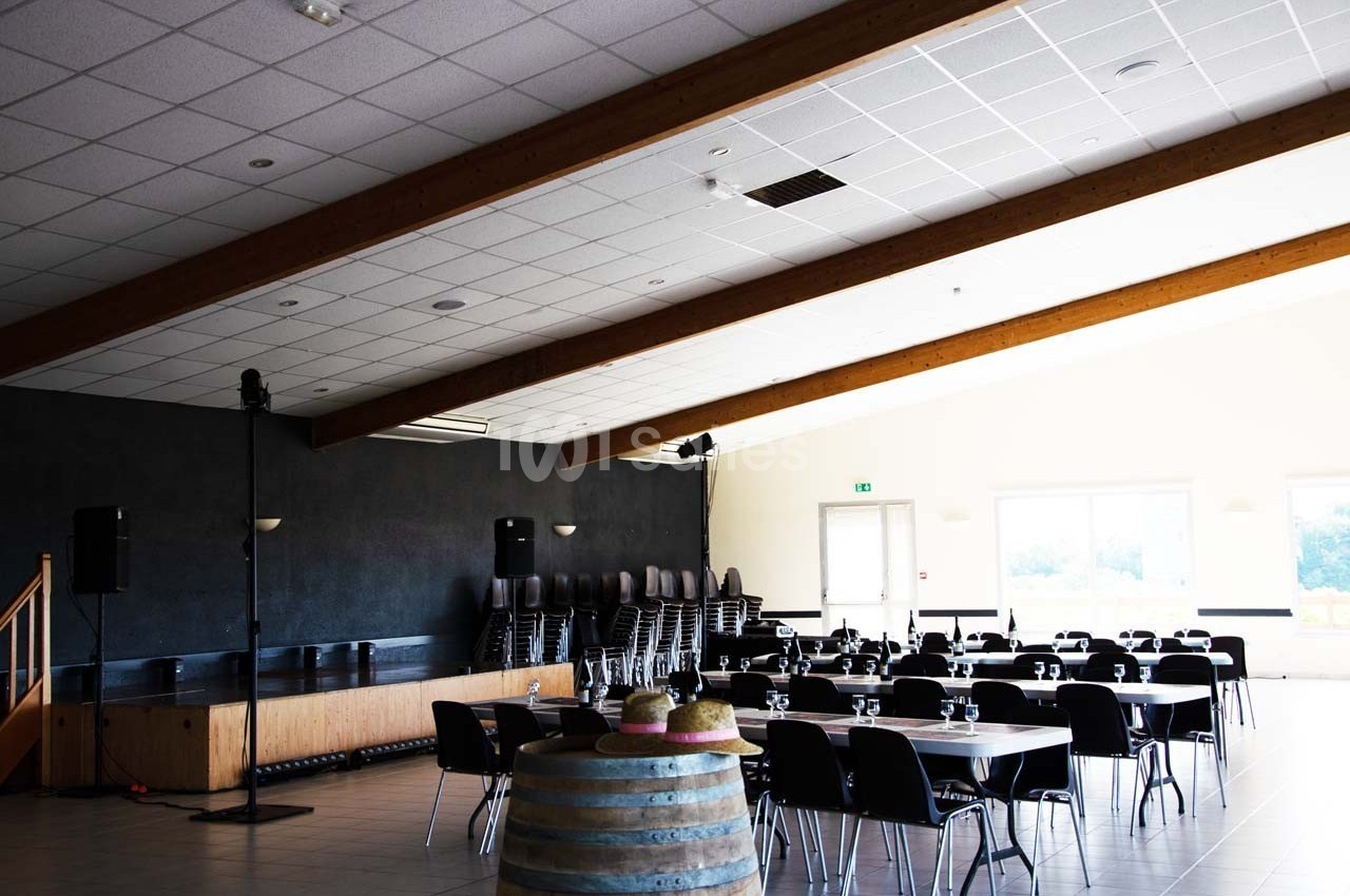 Salle lumineuse avec tables rondes dressées, chaises empilées dans un coin et tonneau décoratif au premier plan.