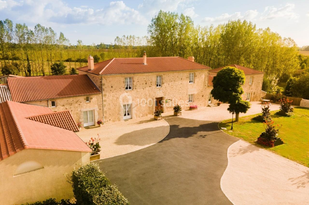 Vue aérienne d'une grande maison en pierre avec toit en tuiles rouges, entourée d'arbres et d'une cour aménagée.