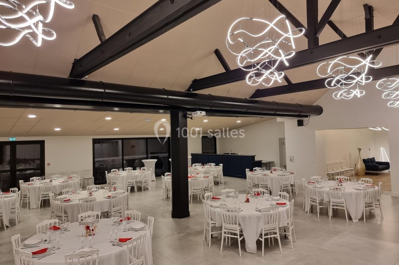 Salle de réception moderne avec tables rondes dressées, chaises blanches et éclairage design suspendu.