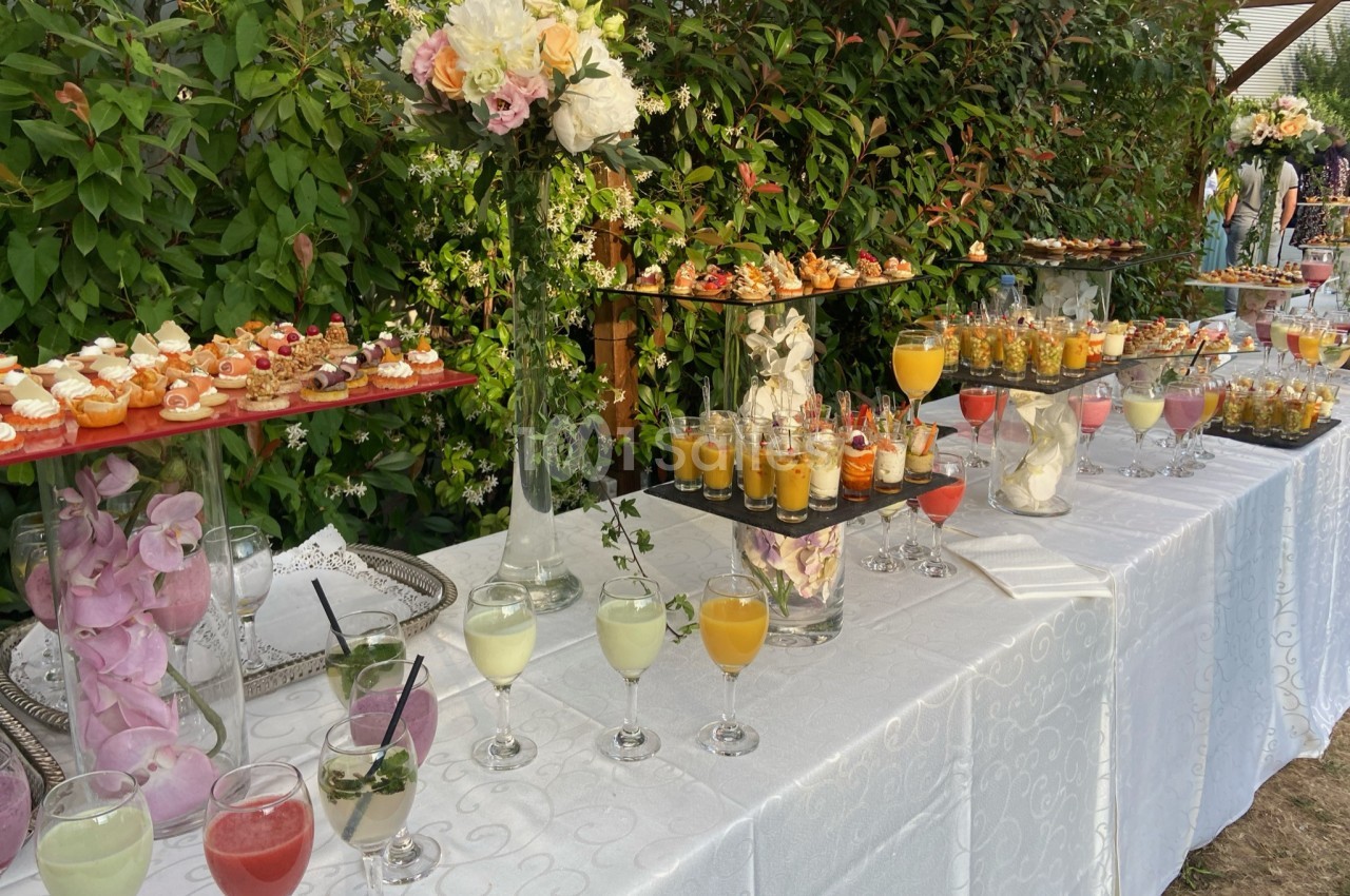Buffet en extérieur avec verrines, cocktails colorés et petits fours, décoré de fleurs et entouré de verdure.