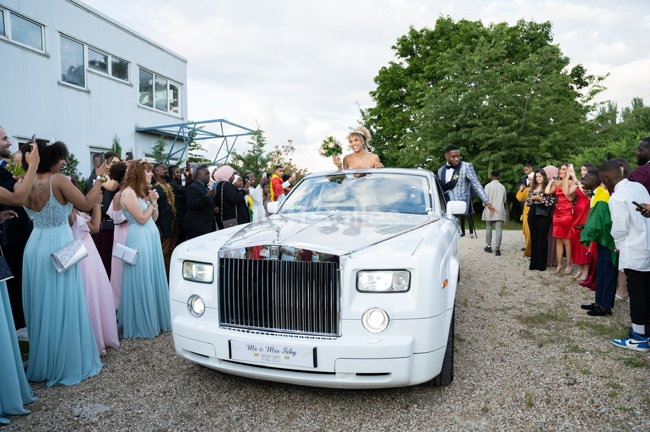 Une mariée et un marié dans une voiture blanche entourés d'invités en tenue de cérémonie.
