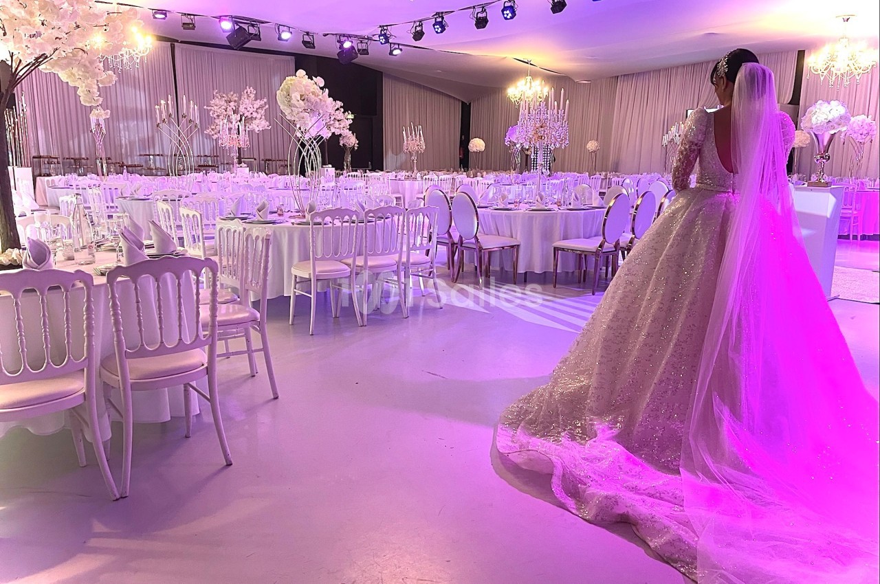 Une mariée en robe blanche observe une salle de réception décorée de fleurs blanches et éclairée de lumières violettes.
