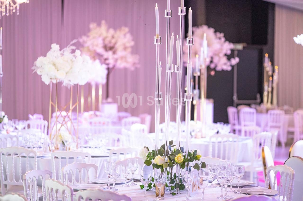 Salle de réception décorée avec des tables dressées, chandeliers modernes et arrangements floraux blancs.