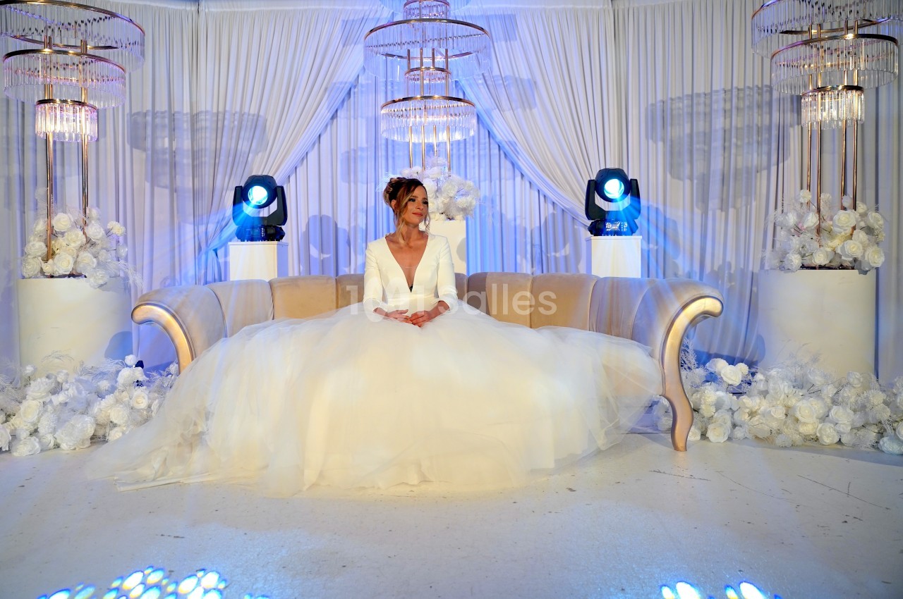 Une femme en robe de mariée blanche est assise sur un canapé beige, entourée de décorations florales et lumineuses.