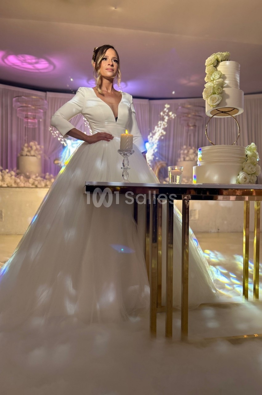 Une femme en robe de mariée blanche se tient près d'une table avec un gâteau décoré de fleurs blanches.