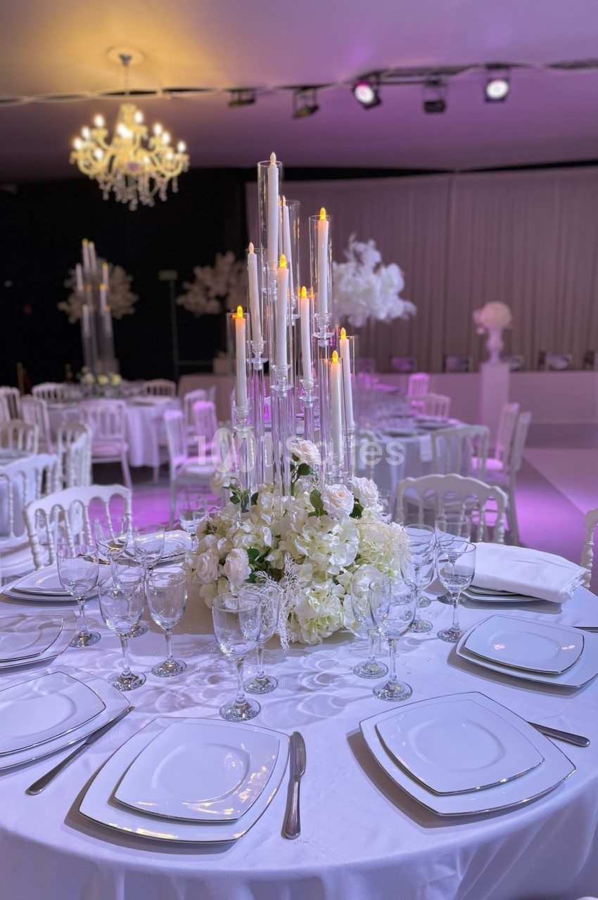 Centre de table avec chandeliers et fleurs blanches, entouré de tables dressées dans une salle de réception élégante.
