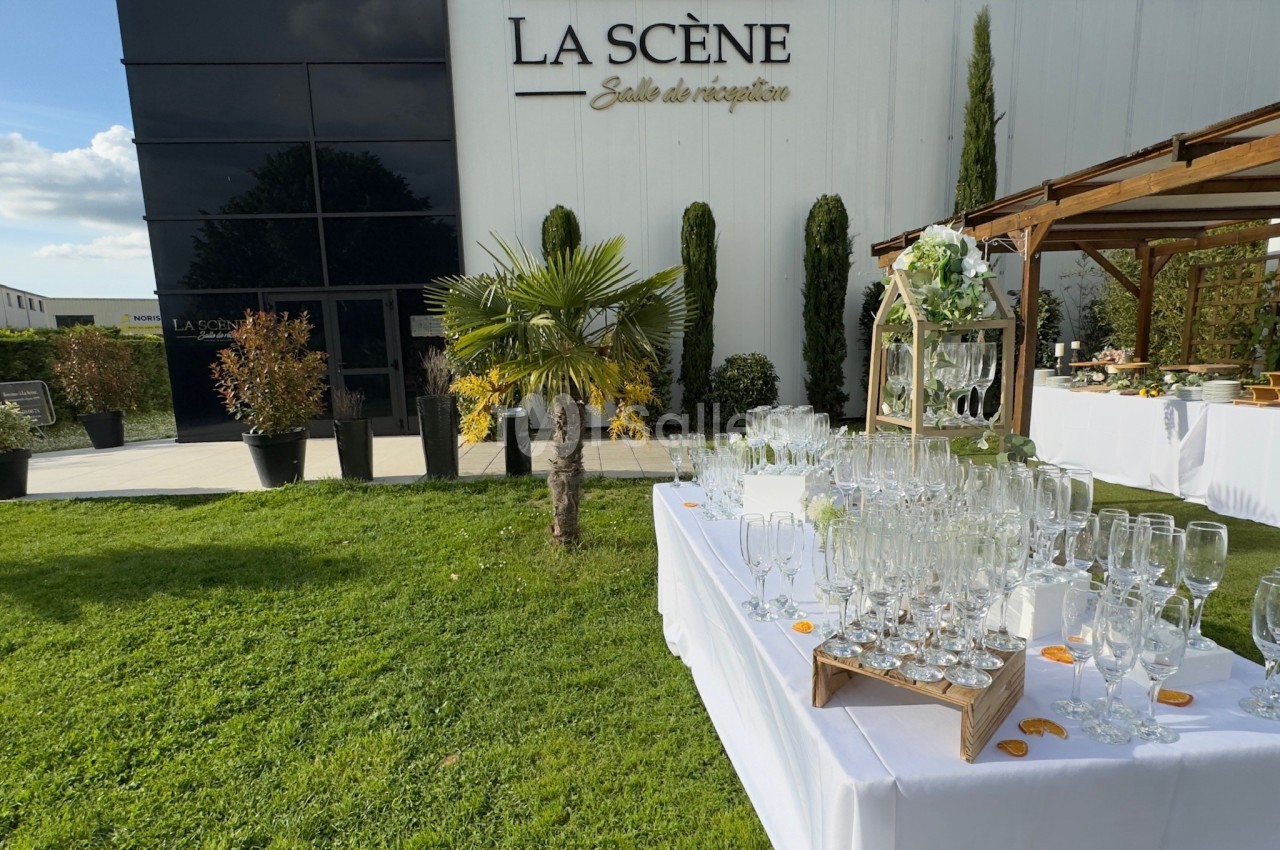 Extérieur d'une salle de réception avec tables dressées, verres et décorations sur une pelouse ensoleillée.