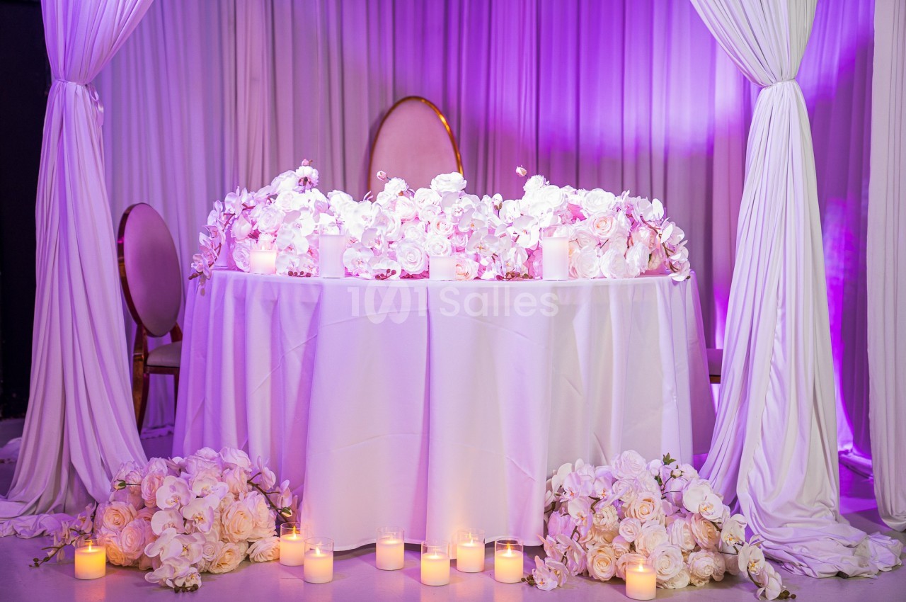 Table décorée de fleurs blanches et roses, entourée de bougies allumées, sous des rideaux blancs éclairés en violet.