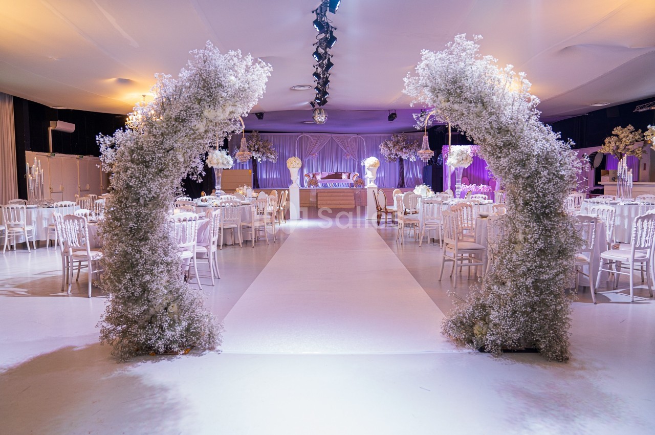 Salle de réception décorée avec des arches fleuries blanches, tables dressées et éclairage tamisé.