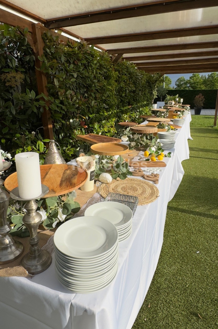 Buffet en extérieur avec nappes blanches, vaisselle, bougies et décorations végétales sur une pelouse.