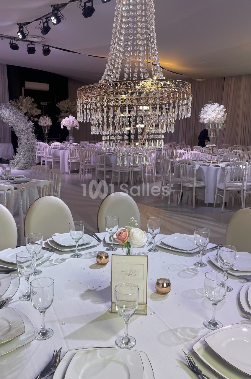 Salle de réception élégante avec tables dressées, grand lustre en cristal et décoration florale blanche.