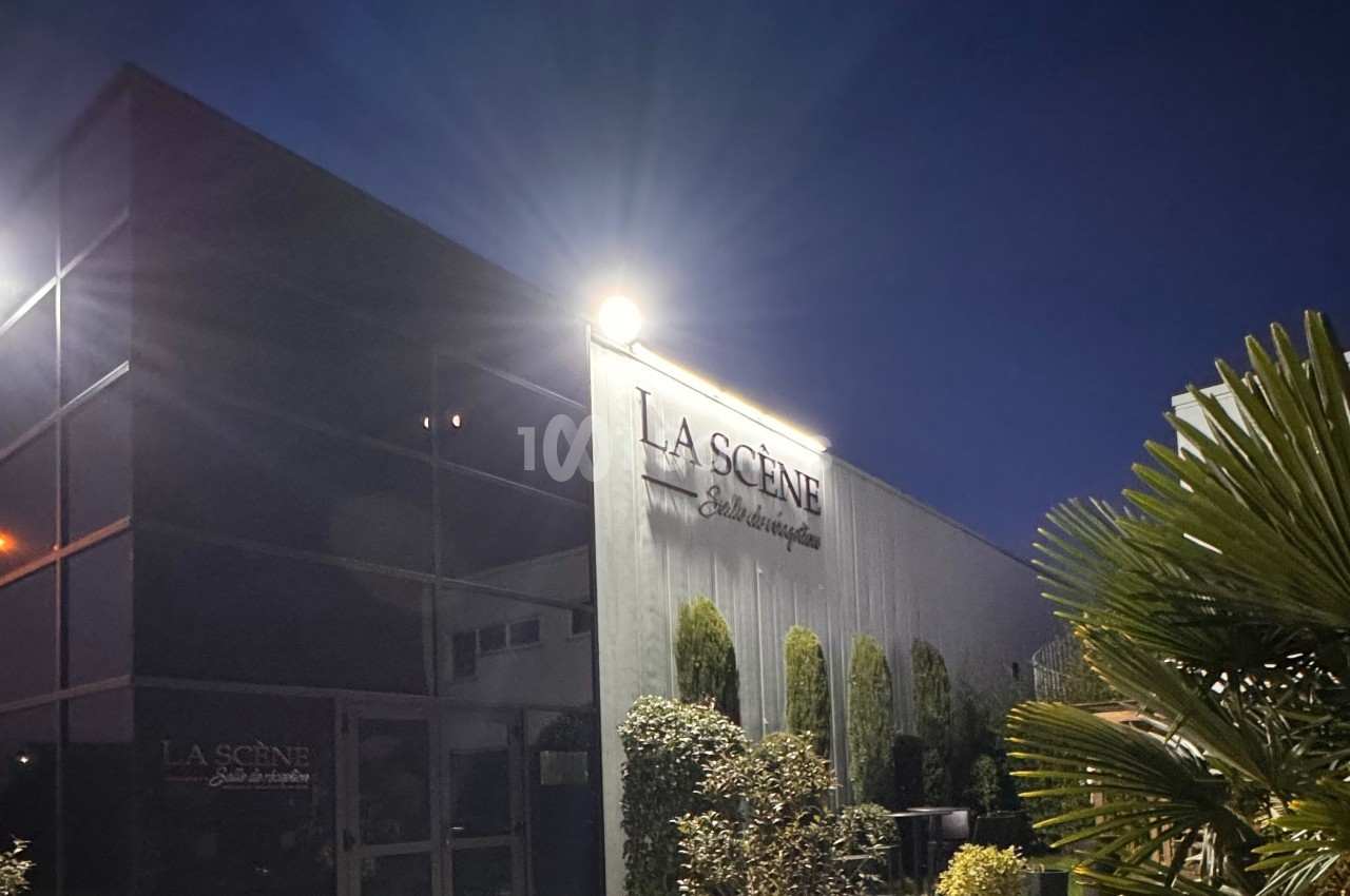 Façade d'un bâtiment moderne éclairé de nuit, avec des plantes et un panneau ’La Scène’ visible.