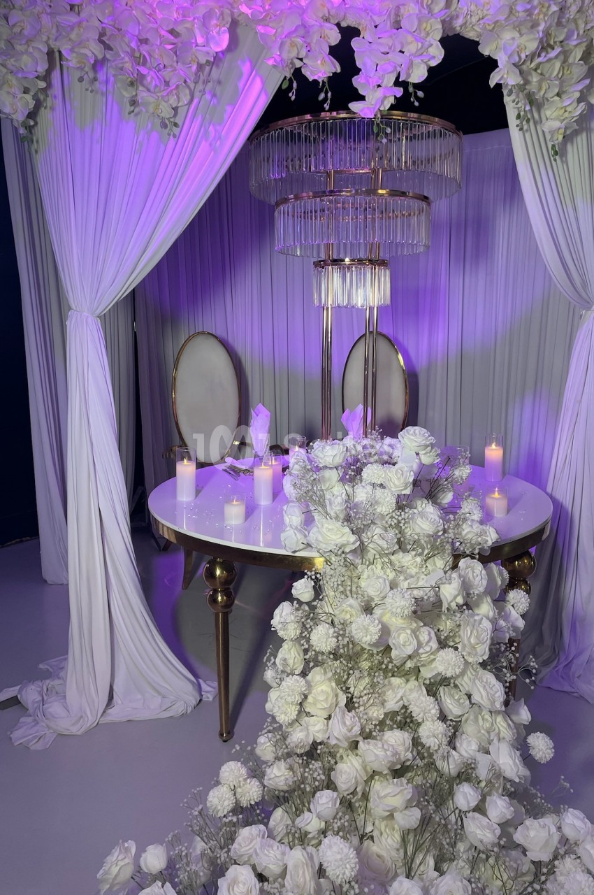 Décoration élégante avec table blanche, chaises ovales, fleurs blanches et rideaux drapés sous un éclairage violet.