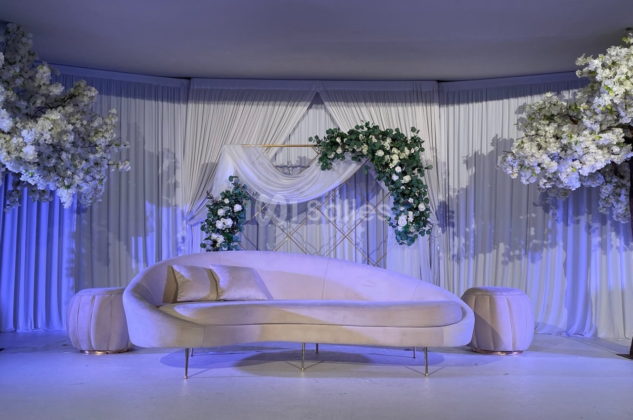 Canapé beige incurvé devant un décor élégant avec rideaux blancs, arches fleuries et arbres ornés de fleurs blanches.