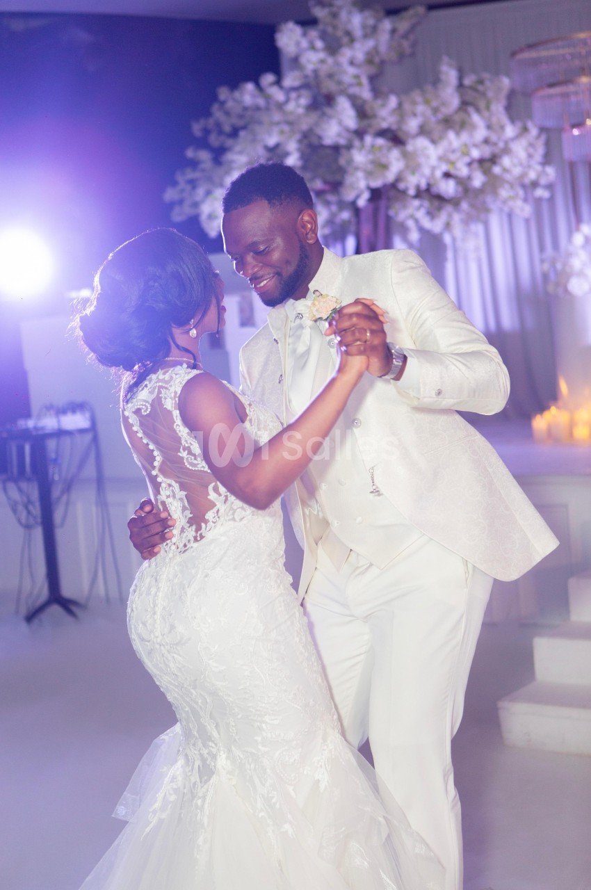 Un couple en tenue de mariage danse dans une salle décorée de fleurs blanches et éclairée par une lumière douce.