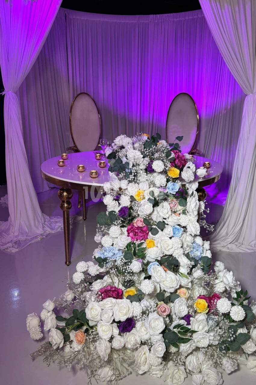 Table décorée avec des chaises élégantes, drapés blancs, éclairage violet et cascade de fleurs multicolores.