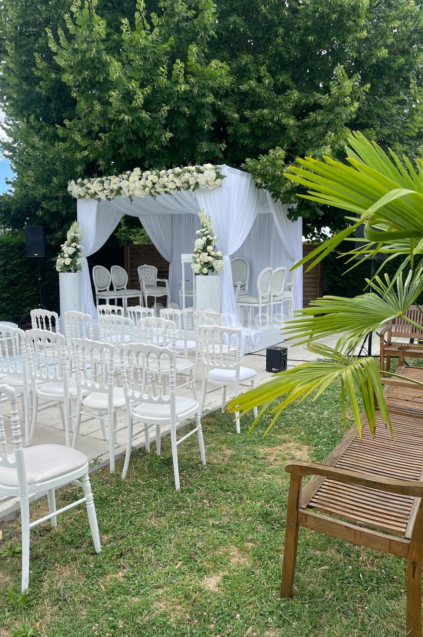 Cérémonie en plein air avec arche fleurie blanche, chaises blanches alignées et bancs en bois dans un jardin verdoyant.