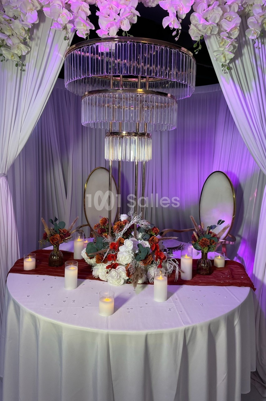 Table décorée avec des fleurs rouges et blanches, bougies allumées et lustre suspendu, entourée de rideaux blancs.