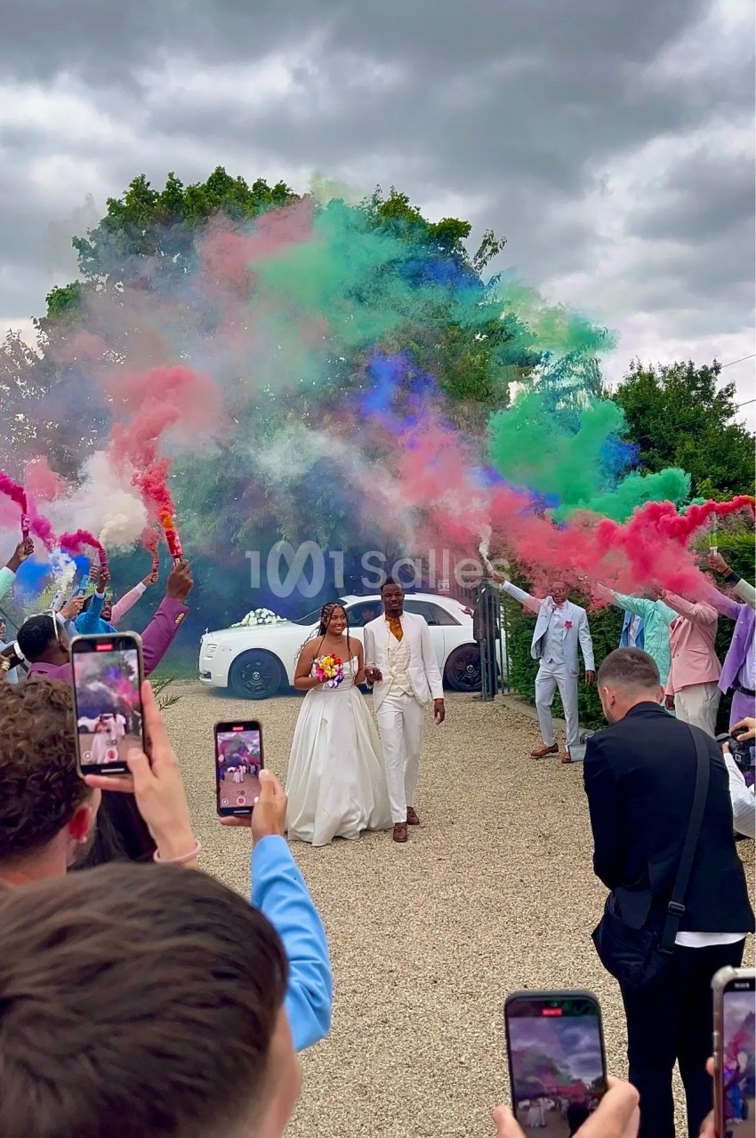Un couple en tenue de mariage marche sous des fumigènes colorés, entouré d'invités prenant des photos.