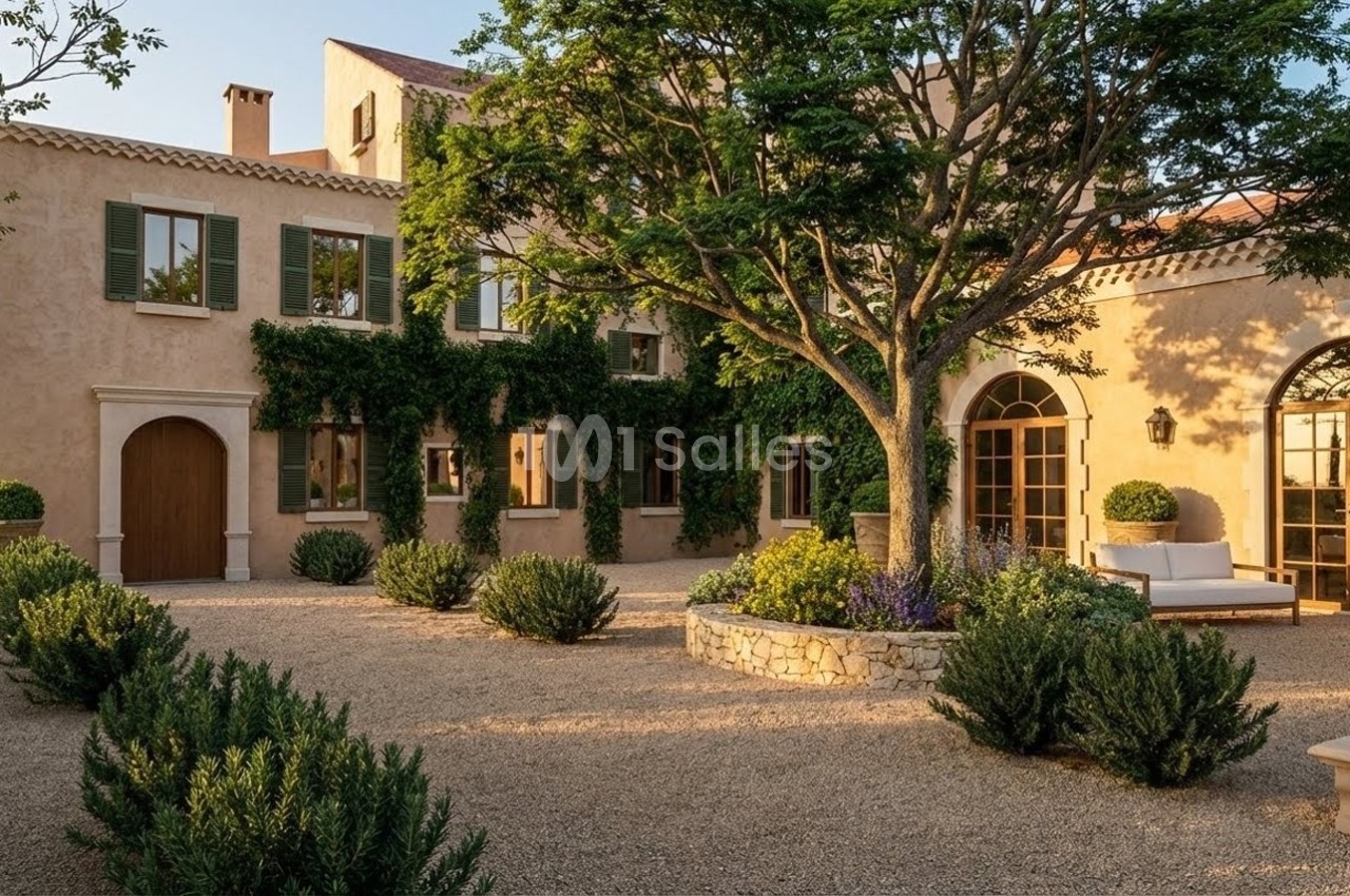 Façade d'une maison en pierre avec volets verts, entourée de végétation et d'une cour en gravier.
