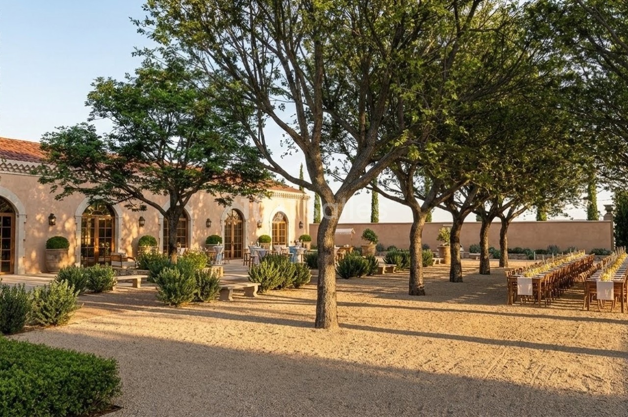 Allée bordée d'arbres menant à une bâtisse en pierre avec tables dressées pour un repas en extérieur.