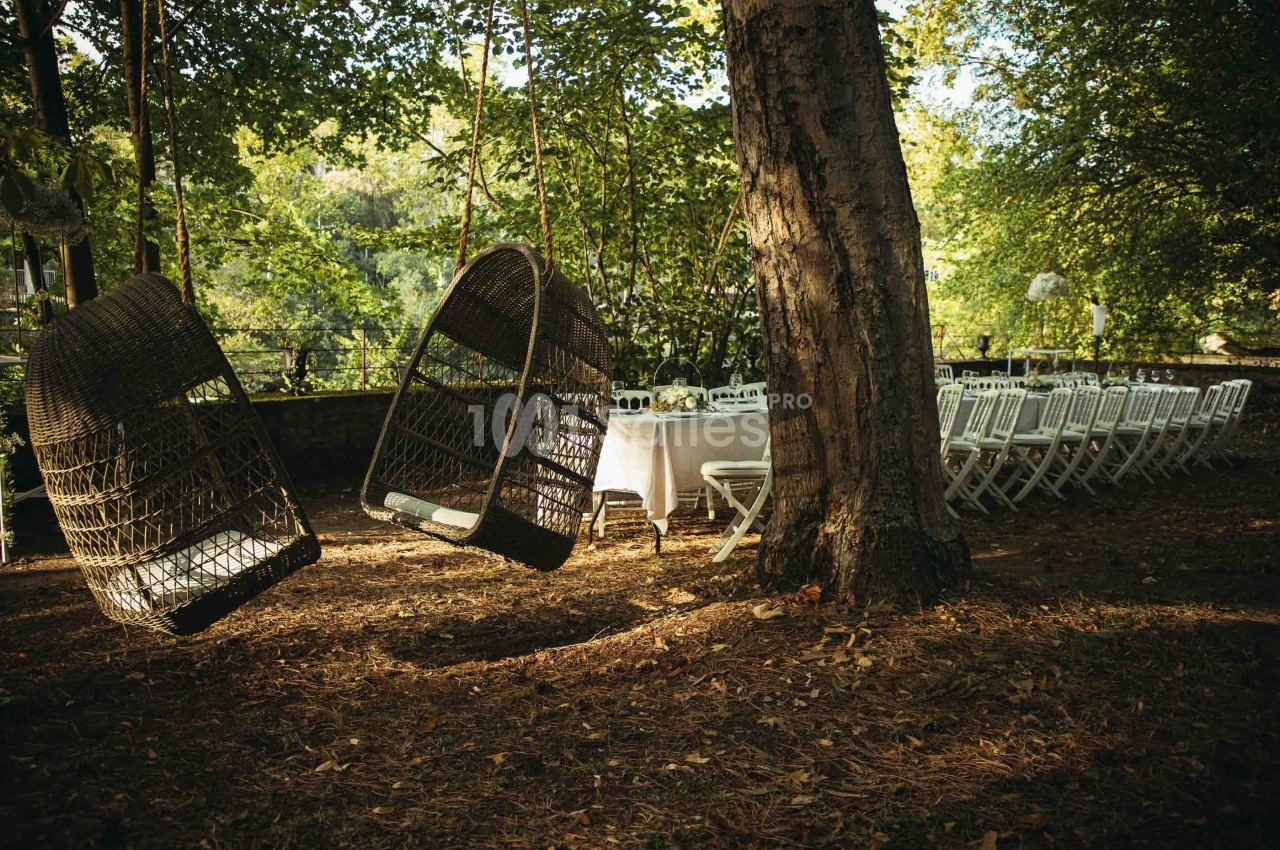 Deux fauteuils suspendus en osier près d'une table dressée avec des chaises blanches dans un jardin ombragé.
