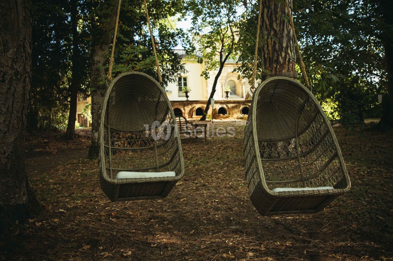 Deux fauteuils suspendus en osier installés sous des arbres dans un jardin, avec une maison en arrière-plan.