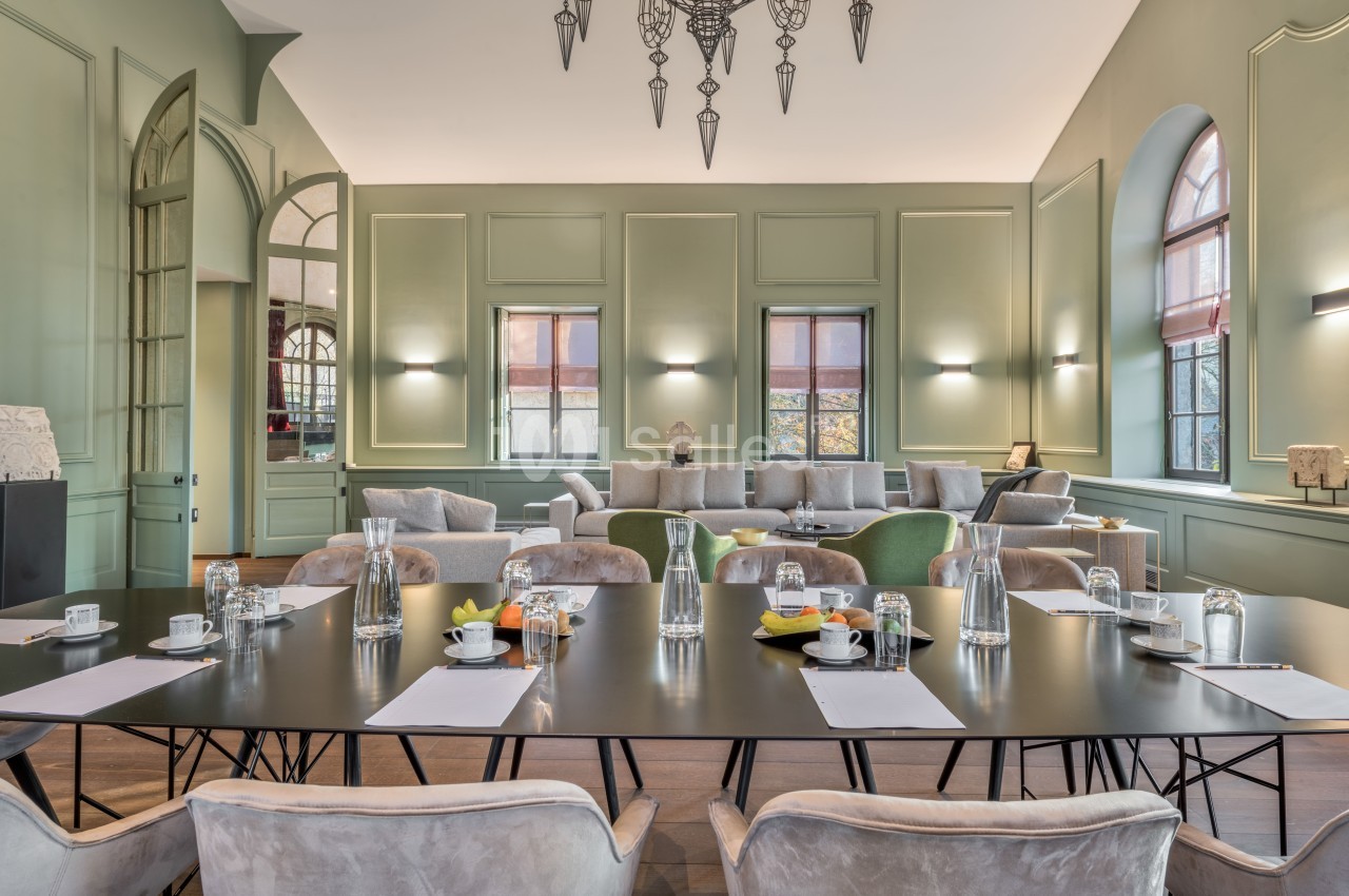 Salle lumineuse avec table de réunion, chaises en velours beige, canapés confortables et murs verts élégants.