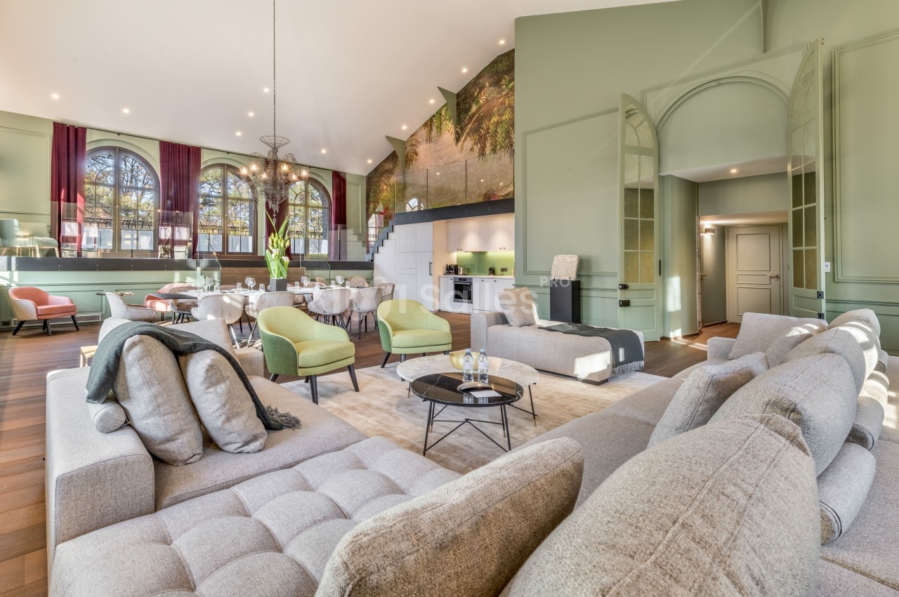 Salon spacieux avec canapé beige, fauteuils verts, grande table à manger, hauts plafonds et mur décoré d'une fresque.