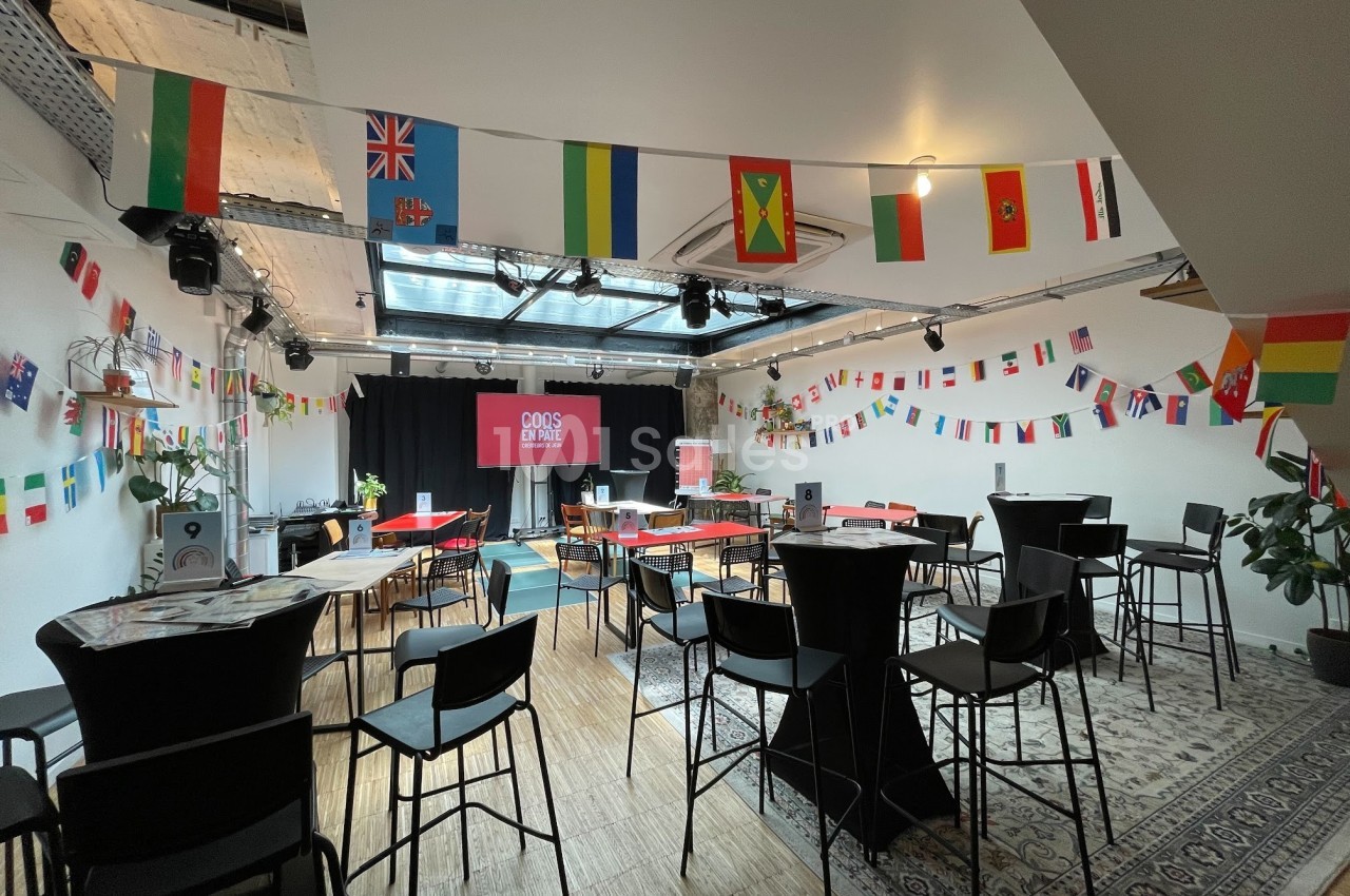 Salle décorée de drapeaux internationaux, aménagée avec des tables hautes et des chaises pour un événement.