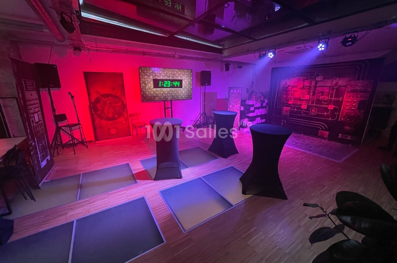 Salle de jeu immersive avec éclairage coloré, écran affichant un compte à rebours et tables hautes noires.
