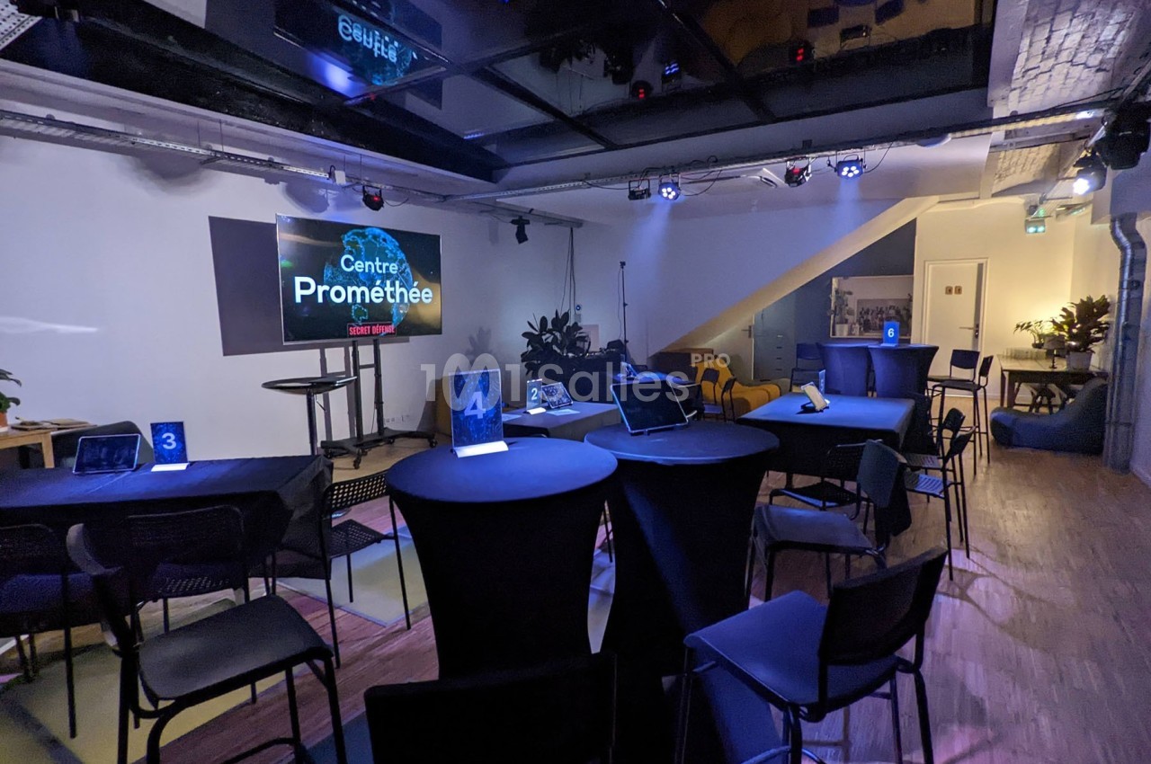 Salle de conférence moderne avec tables hautes, chaises, écrans et éclairage tamisé.