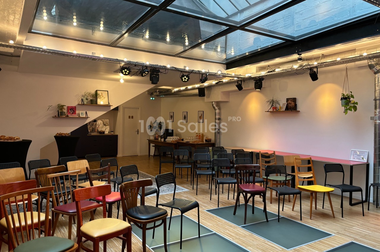 Salle lumineuse avec verrière, chaises disposées pour un événement, décorations murales et tables avec collations.