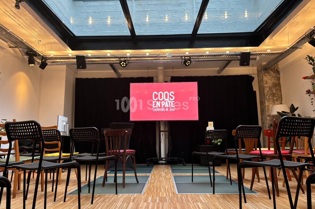 Salle lumineuse avec des chaises disposées face à un écran affichant ’Coqs en Pâte’ sous une verrière.