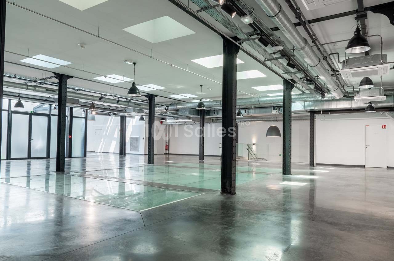 Intérieur d'un espace industriel rénové avec sol en béton, verrières, poutres métalliques et éclairage moderne.